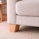 Beige Upholstered Sofa with a Wooden Block Leg on a Light Beige Rug Near a Wooden Table Edge in the Background