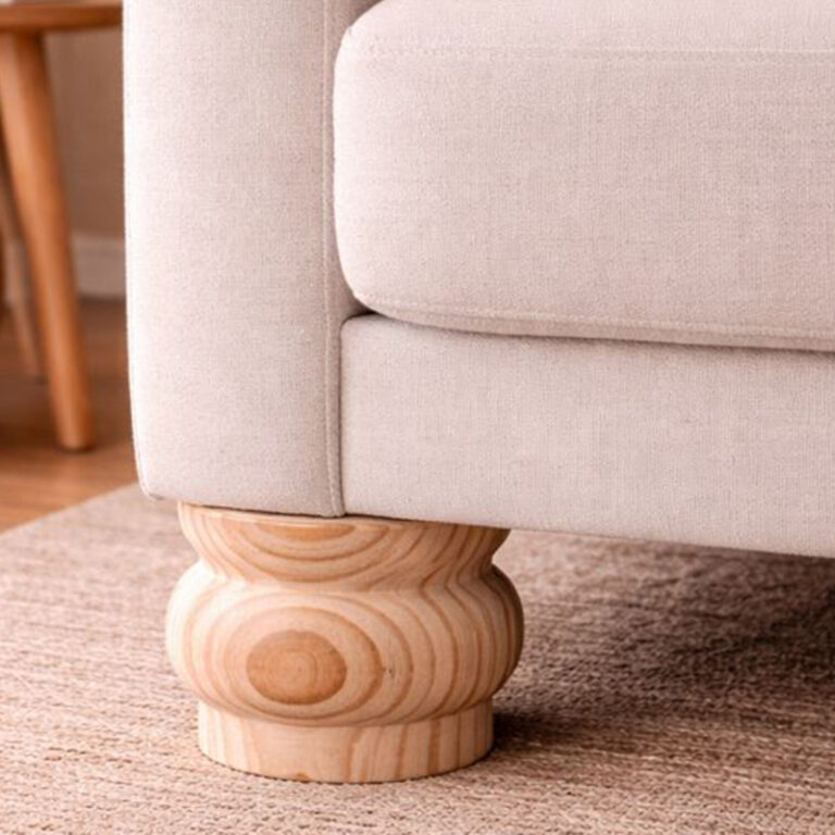 Close-up of a beige upholstered sofa with a rounded, turned wooden leg sitting on a light brown rug.