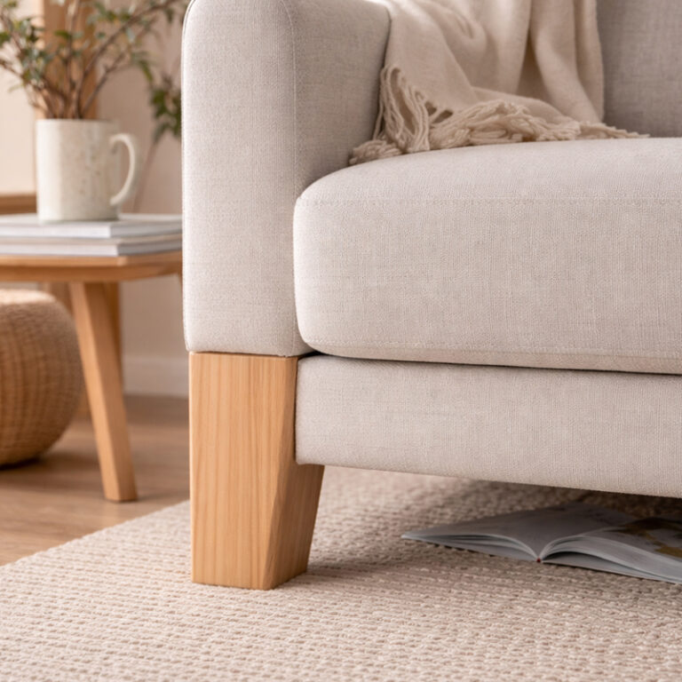 Beige sofa with wooden legs in a cozy living room; a magazine under the seat and a side table with a mug in the background.