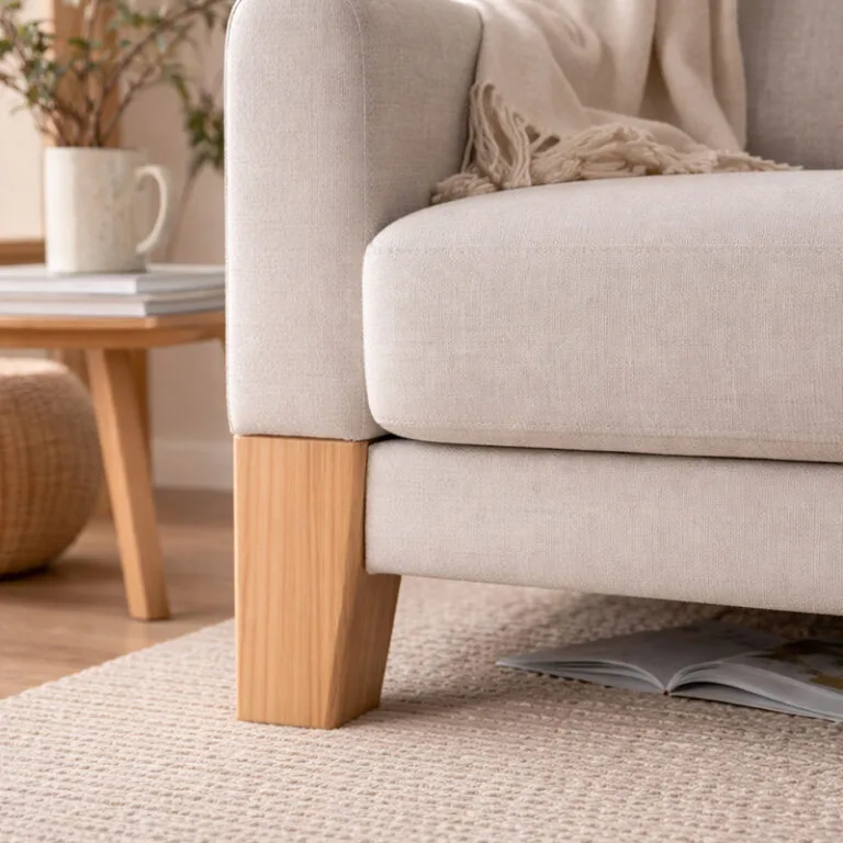 Beige sofa with wooden legs in a cozy living room; a magazine under the seat and a side table with a mug in the background.