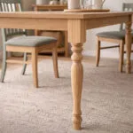 Close up of a Wooden Dining Table Leg with Turned Details in a Light Cozy Dining Room with Upholstered Chairs and a Woven Rug