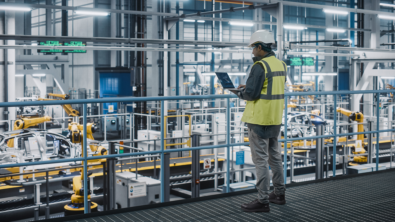 Wright-Glide-Sneaker-Lifestyle | Securadeal Factory Floor with Yellow Robotic Arms and a Worker in a High visibility Vest and Hard Hat Inspecting a Tablet Screen in Front of Equipment