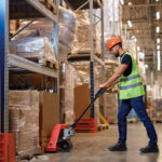 Wright-KPU-Sneaker-Lifestyle | Securadeal Warehouse Worker in a Neon Vest and Orange Hard Hat Operating a Pallet Jack Beside Tall Shelves Loaded with Wrapped Pallets