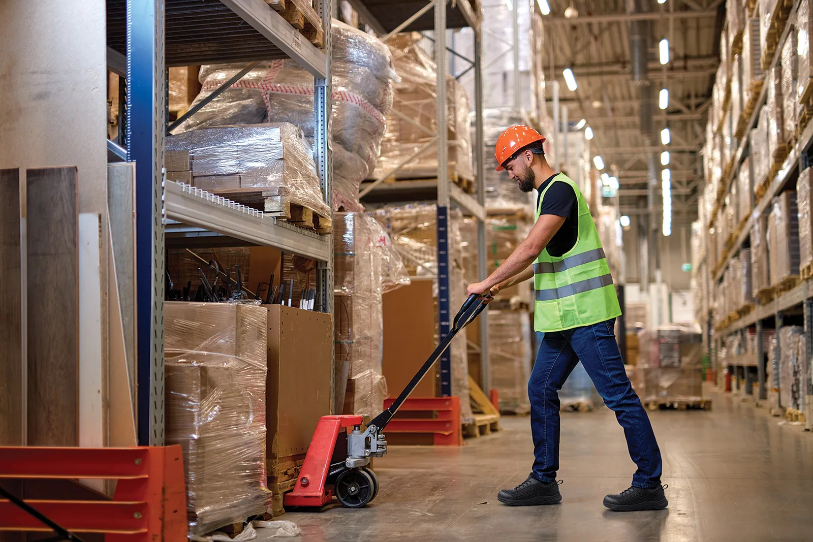 Warehouse Worker in a Neon Vest and Orange Hard Hat Operating a Pallet Jack Beside Tall Shelves Loaded with Wrapped Pallets