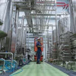 Wright-Sprinter-Sneaker-Lifestyle | Securadeal Maintenance Worker in Blue Overalls and Hard Hat Stands on an Orange Lift Among Stainless Steel Process Pipes and Tanks in a Factory Setting
