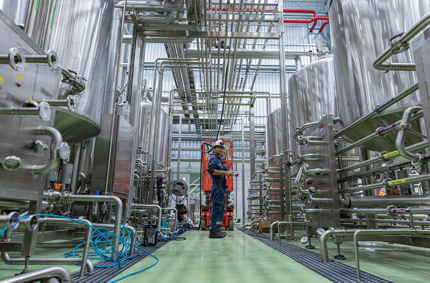 Maintenance Worker in Blue Overalls and Hard Hat Stands on an Orange Lift Among Stainless Steel Process Pipes and Tanks in a Factory Setting