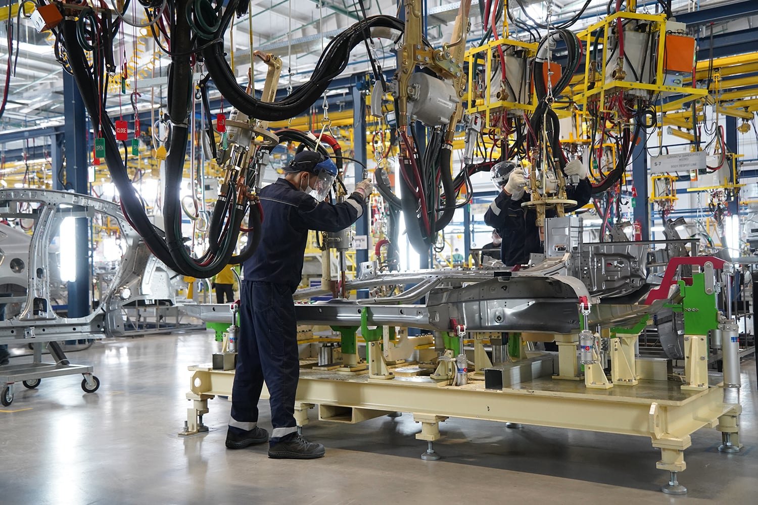 Wright-Supreme-Sneaker-Lifestyle | Securadeal Automotive factory scene with workers in protective gear assembling a car frame on a sunny-green platform, surrounded by complex overhead machinery and cables.