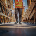 Wright-Dealer-Chelsea-Lifestyle | Securadeal Construction Worker in an Orange Safety Vest and Hard Hat Walks Between Rusted Metal Structures on a Scaffolded Site