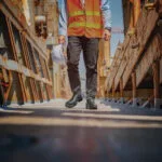 Wright-Dealer-Chelsea-Lifestyle | Securadeal Construction Worker in an Orange Safety Vest and Hard Hat Walks Between Rusted Metal Structures on a Scaffolded Site