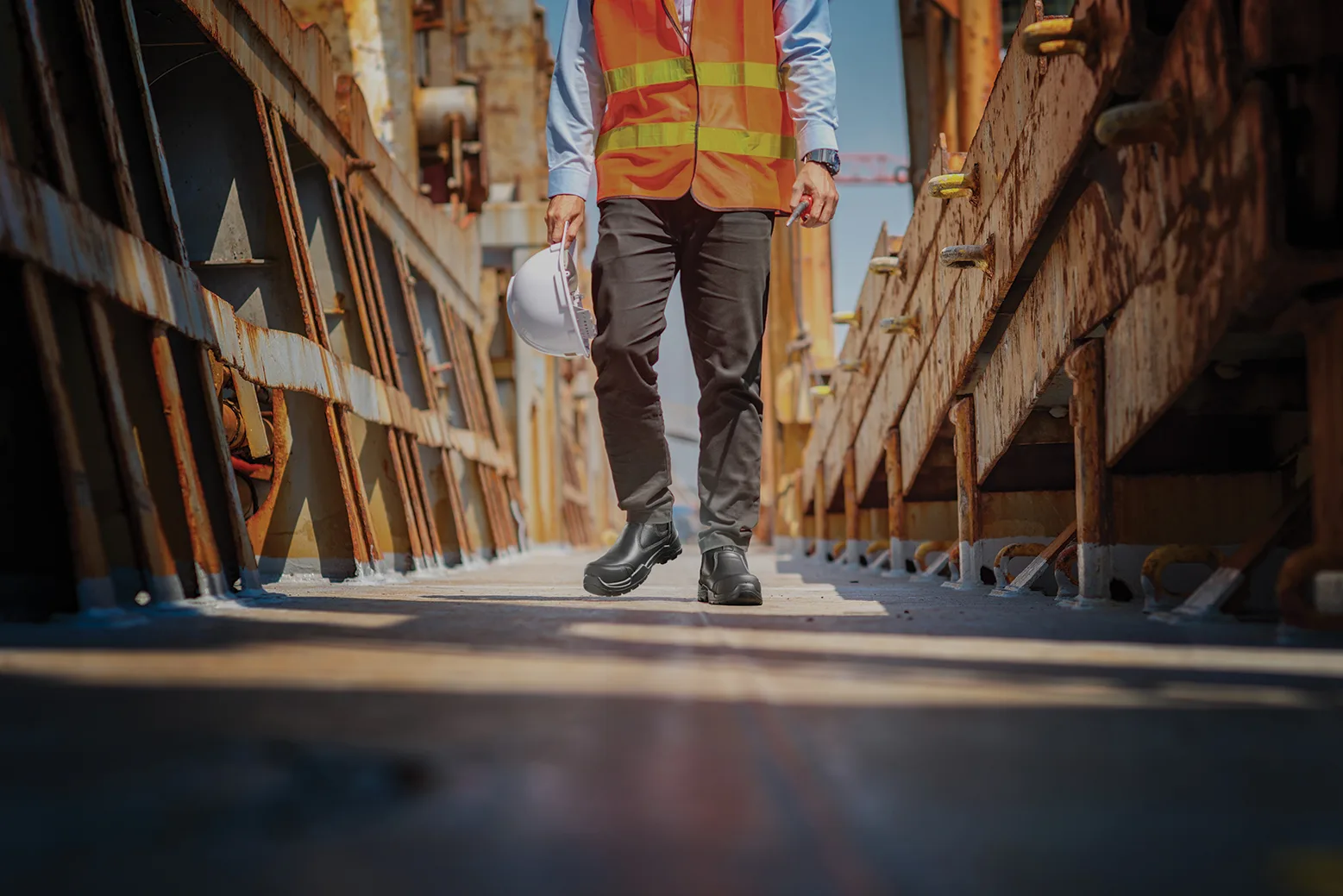 Construction Worker in an Orange Safety Vest and Hard Hat Walks Between Rusted Metal Structures on a Scaffolded Site