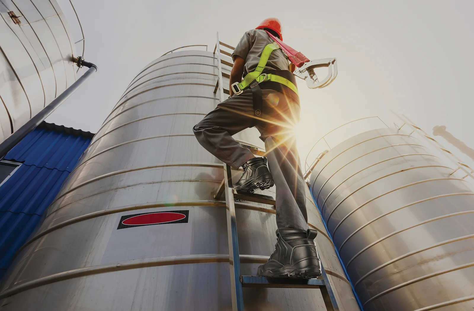 Construction Worker Wearing Hard Hat and Safety Harness Climbs a Ladder Beside Large Silver Silos