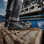 Wright-Hiker-Edge-Lifestyle | Securadeal Close up of a Workers Black Safety Boots on a Rusty Metal Platform Beside Blue Industrial Machinery