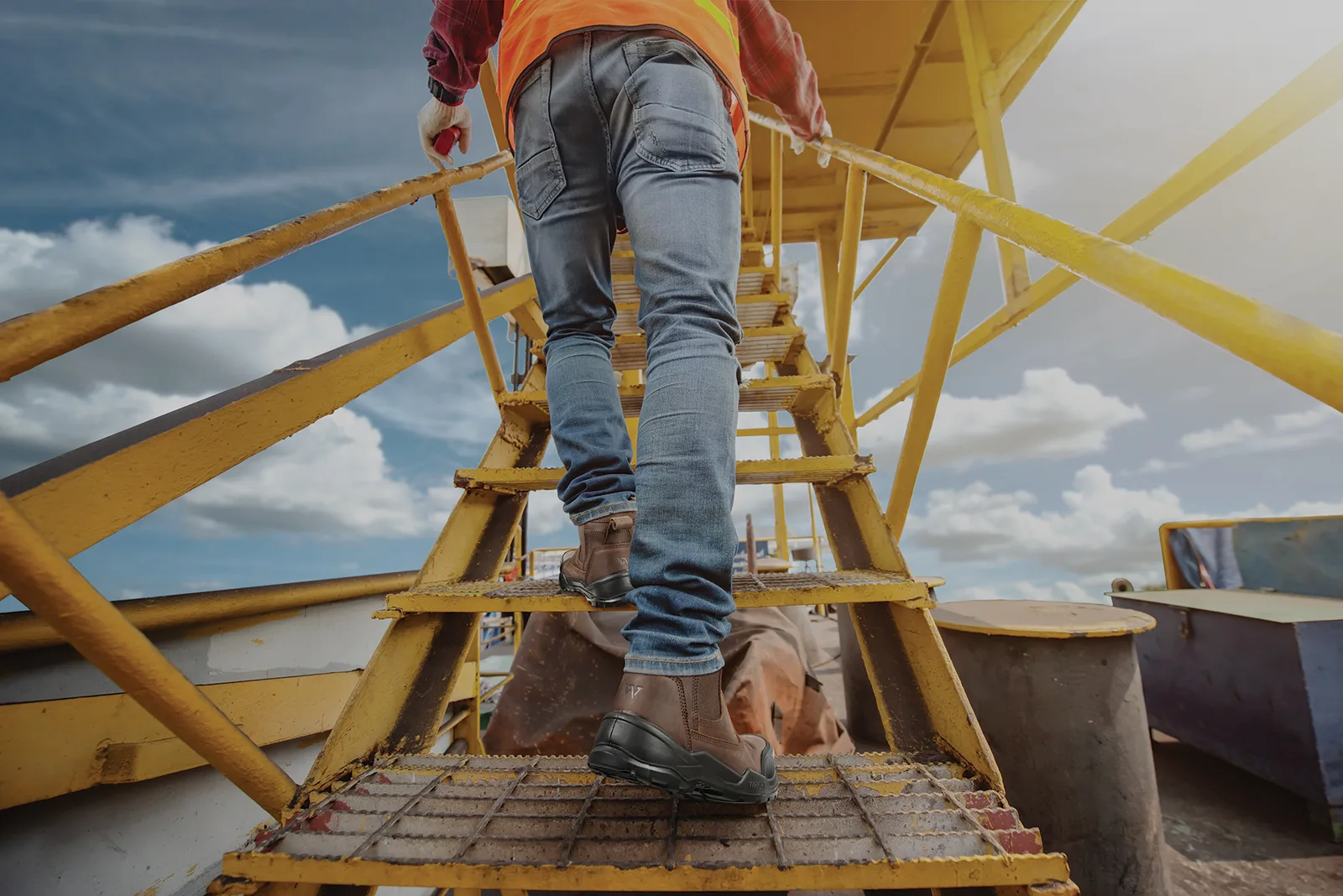 Construction Worker in an Orange Safety Vest Climbs a Yellow Metal Staircase on an Outdoor Site