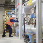 Worker in Orange Safety Vest and White Helmet Inspecting a Large Industrial Piping System in a Factory Environment