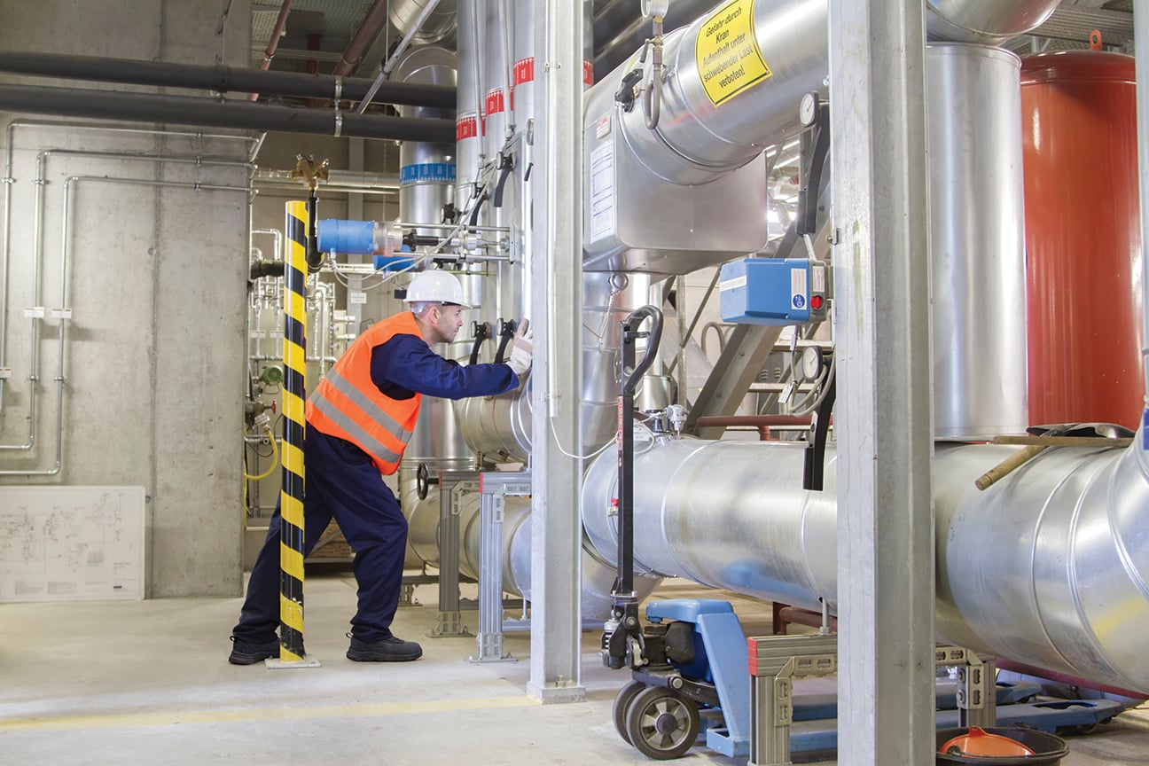 Wright-PU-Tek-Sneaker-Lifestyle | Securadeal Worker in orange safety vest and white helmet inspecting a large industrial piping system in a factory environment.