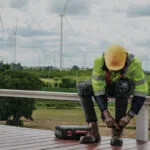 Wright-Sierra-Brown-Lifestyle | Securadeal Worker in High visibility Jacket and Hard Hat Kneels on a Railing with Tools Wind Turbines in a Rural Field Behind Him