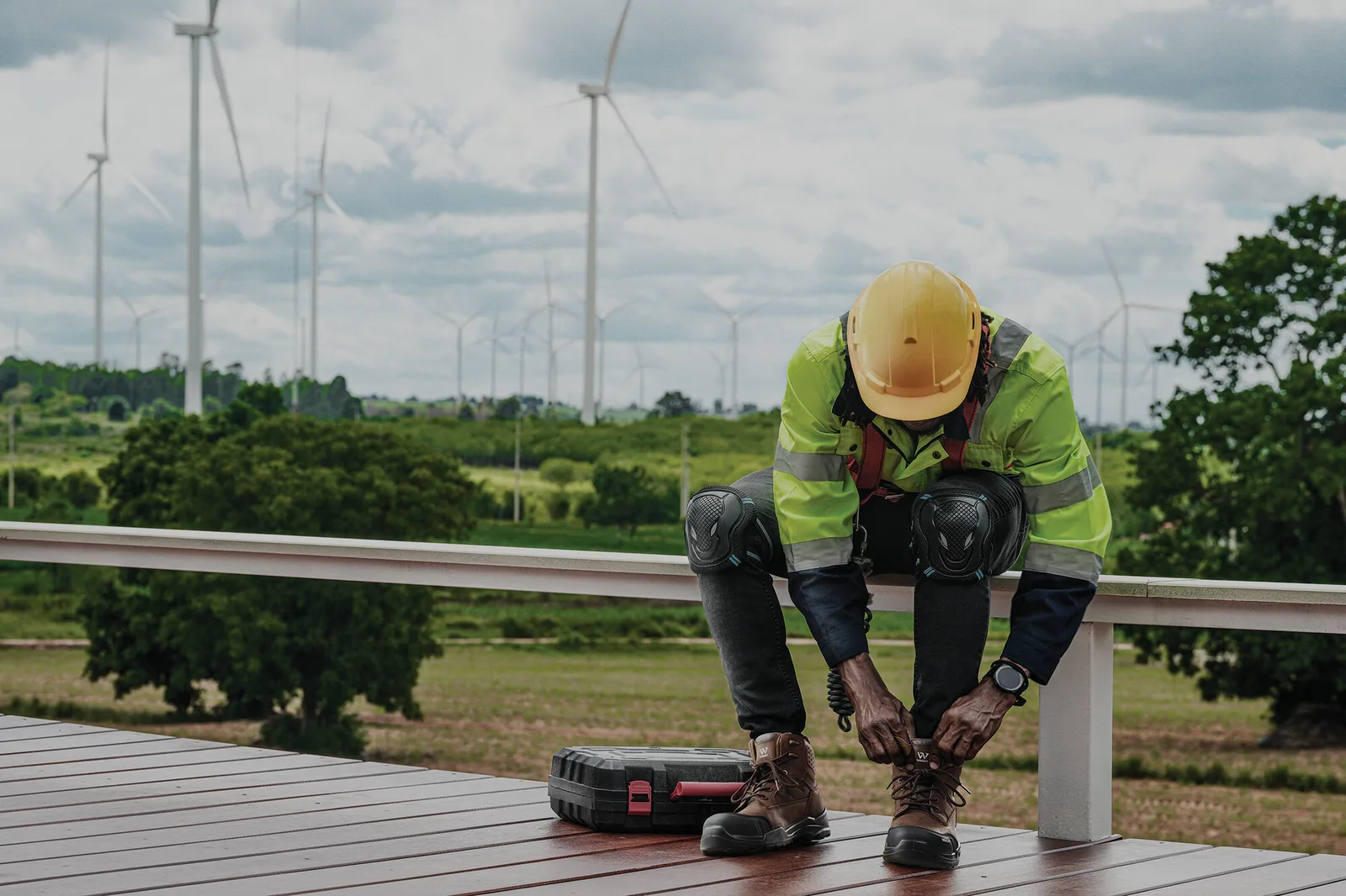 Worker in High visibility Jacket and Hard Hat Kneels on a Railing with Tools Wind Turbines in a Rural Field Behind Him