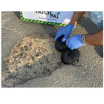 3 | Securadeal Worker in Blue Gloves Fills a Pothole with Black Repair Material in a Road Beside a Faithpal Pothole Solution Sign