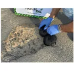 3 | Securadeal Worker in Blue Gloves Fills a Pothole with Black Repair Material in a Road Beside a Faithpal Pothole Solution Sign